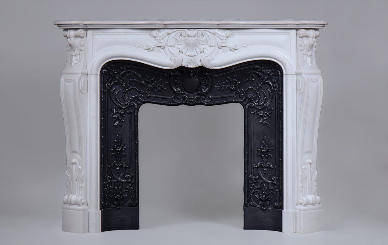 «Madame Du Barry» est une cheminée de style Louis XV en marbre Blanc de Carrare d’une rare opulence.
Les formes généreuses sont richement décorées de motifs végétaux finement sculptés. Le centre du bandeau de cette cheminée sur mesure est orné d’une large coquille fleurie, motif que nous retrouvons sur les têtes de jambages. Ces derniers, aux formes galbées, dessinent un enroulement dans leur partie haute.
