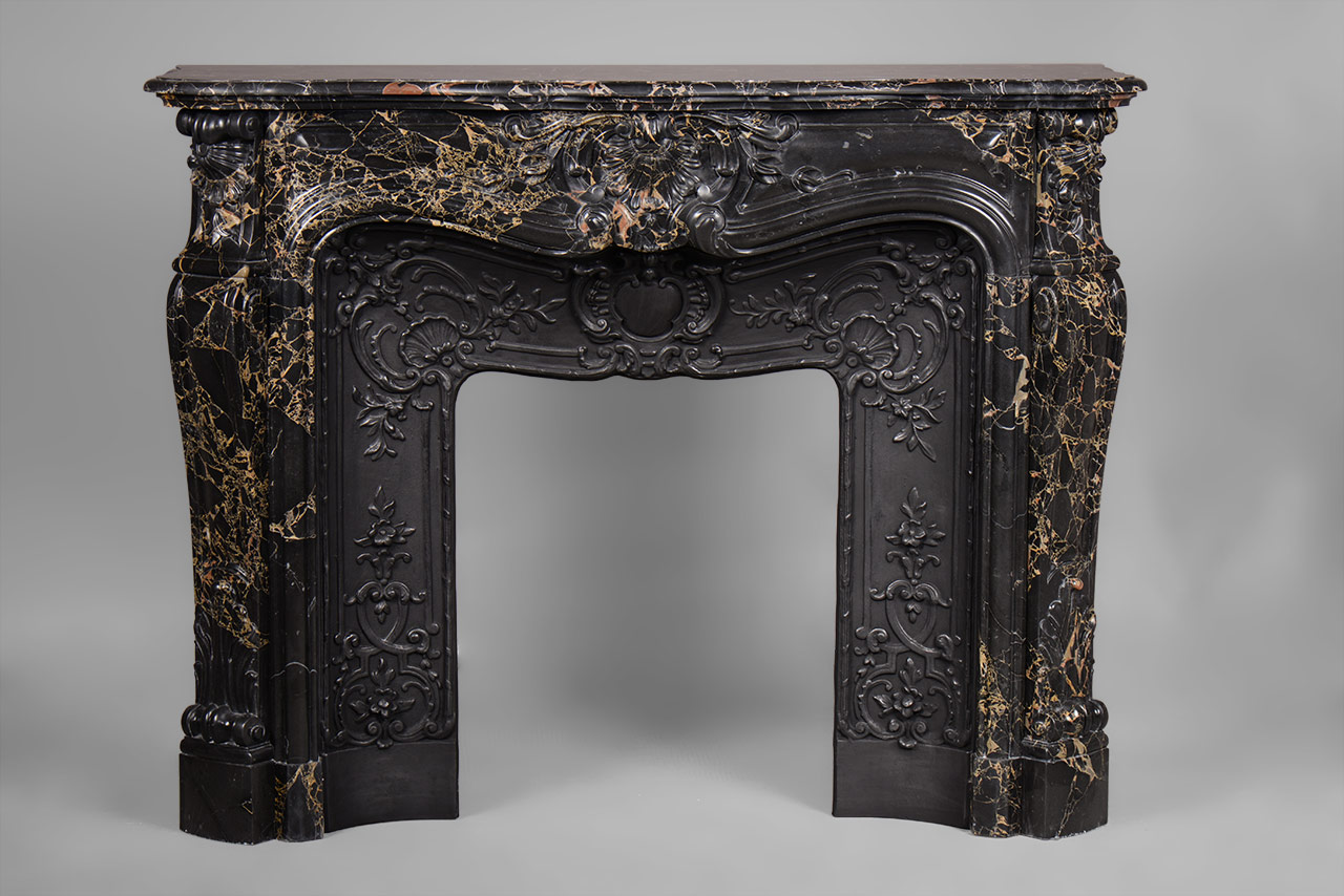 «Madame Du Barry» est une cheminée de style Louis XV en marbre noir Portoro d’une rare opulence.
Les formes généreuses sont richement décorées de motifs végétaux finement sculptés. Le centre du bandeau de cette cheminée sur mesure est orné d’une large coquille fleurie, motif que nous retrouvons sur les têtes de jambages. Ces derniers, aux formes galbées, dessinent un enroulement dans leur partie haute.
